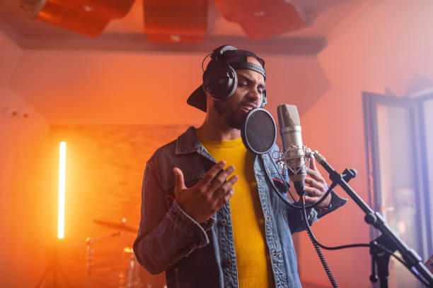 A portrait of a young adult black male RnB singer passionately singing into a microphone and moving his hands. The attractive singer has a beard and wears a baseball cap. He is attractive and is dressed in casual cool clothes. In the background there is a cool orange light illumating the recording studio and some drums.